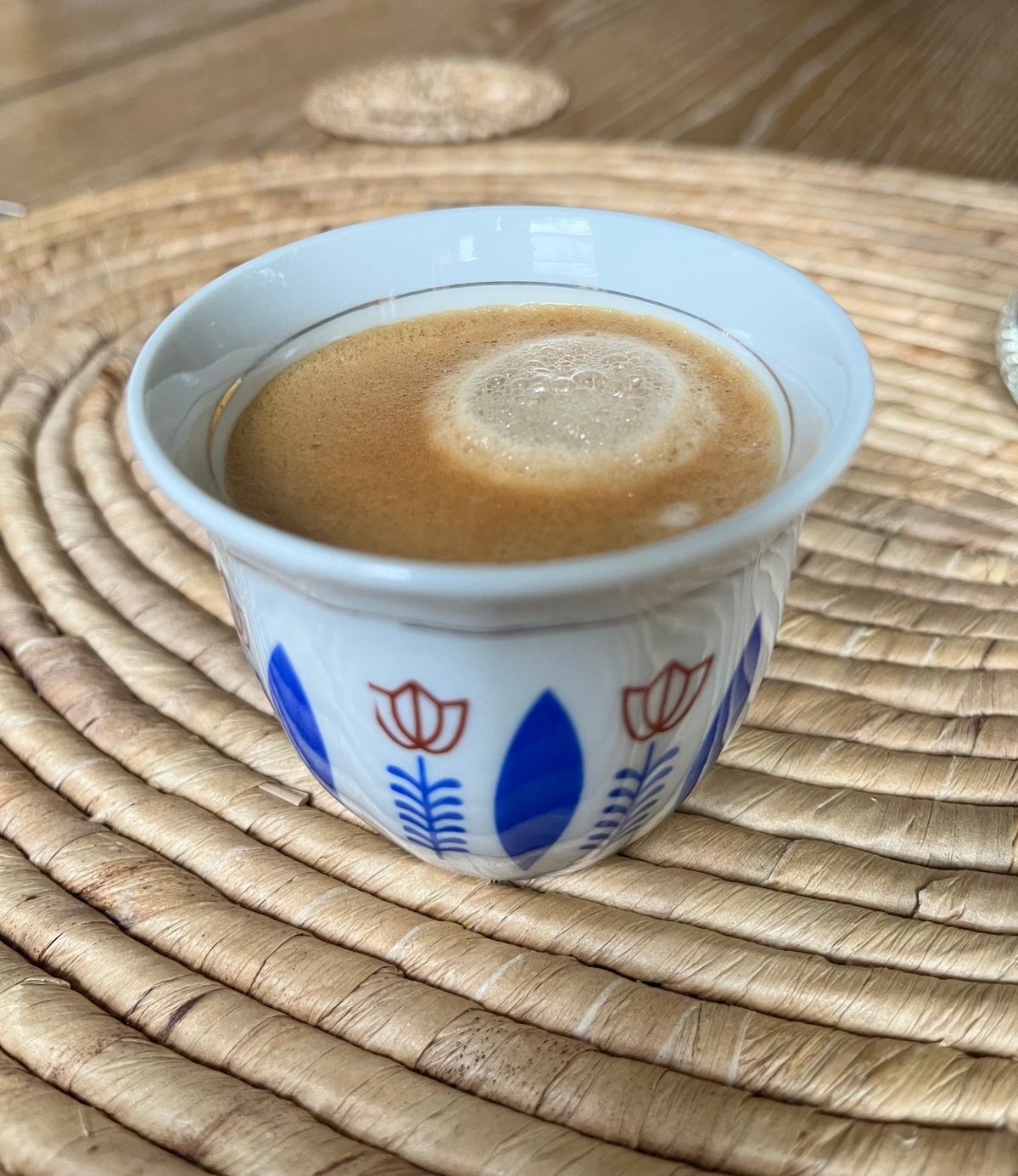 A small handleless cup of frothy coffee painted with blue leaves and red tulips, on a woven placemat