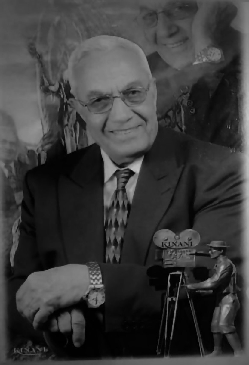 Black-and-white portrait of Marcel Kinani in a suit and tie, smiling, beside a small sculpture of a vintage movie camera bearing the Kinani name