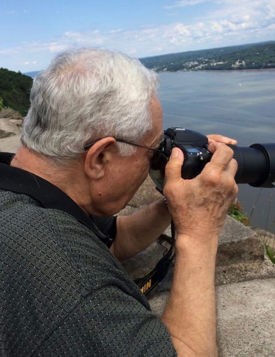 Marcel Kinani, white-haired, looking through a camera with a long lens toward a wide river and wooded hills